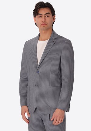 Uomo che indossa una giacca da completo grigia a righe sottili e pantaloni abbinati sopra una camicia bianca, in piedi su uno sfondo chiaro e uniforme.