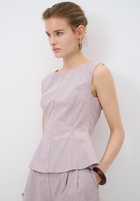 Top peplum senza maniche di color lilla con scollo a barca, tessuto testurizzato e dettagli delle cuciture. Indossato con un bracciale marrone, sullo sfondo neutro.