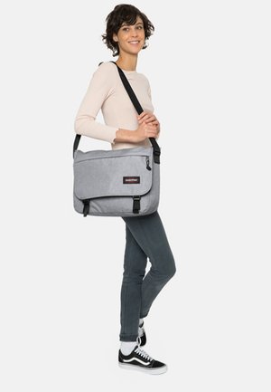 Sac messager gris avec rabat et fermeture à boucle, fabriqué en tissu durable. Il dispose d'une bandoulière et d'un petit patch logo.