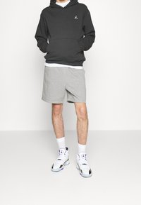 Sudadera negra con un bolsillo frontal y logo, pantalones cortos grises, calcetines blancos y zapatillas blancas con detalles en negro y morado. Atuendo atlético casual.