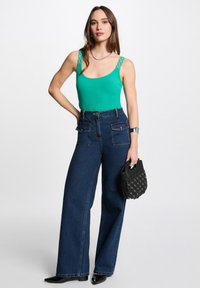 Green ribbed bodysuit with decorative straps, paired with high-waisted, wide-leg dark blue denim jeans. Black handbag with grommet details.