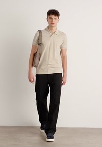 Beige polo skjorte med korte ermer, svarte bukser og marineblå sneakers. Modellen bærer en lys brun skulderveske. Enkel, minimalistisk design.
