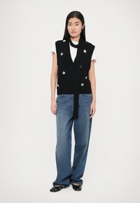 Chaleco negro de punto con botones plateados, superpuesto sobre una camiseta blanca, combinado con vaqueros azules holgados y zapatos negros. Textura suave, diseño casual.