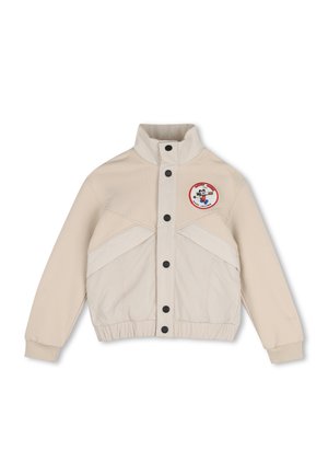 Veste beige avec col haut et boutons noirs, avec un petit patch circulaire représentant un personnage de dessin animé en haut à gauche.
