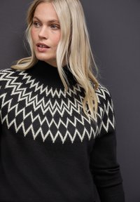 Czarny dzianinowy sweter z białym wzorem w zygzaki na klatce piersiowej i wysokim, prążkowanym kołnierzem. Teksturowany, średniej wagi materiał.