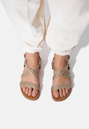 Pies con sandalias de tiras trenzadas color marrón claro con hebillas, combinadas con pantalones blancos con puños sobre fondo blanco.