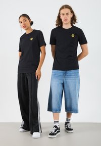 T-shirts en coton noirs avec un petit logo jaune, portés avec des pantalons de jogging noirs et des shorts en denim bleu. Les deux portent des baskets noires avec des accents blancs.