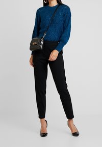 Pull tricoté bleu sarcelle avec motif en losanges texturés, pantalon noir taillé, petit sac bandoulière noir, et escarpins noirs à bout pointu.