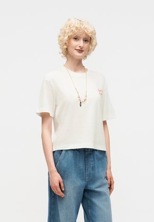 Jeune personne aux cheveux blonds courts et bouclés, portant un t-shirt blanc, un jean bleu et un long collier, debout devant un fond uni.