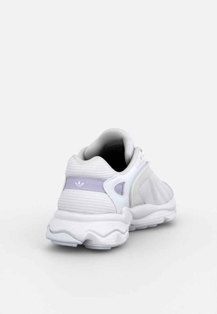 Sneaker bianco sportivo con upper in materiale sintetico testurizzato, suola curva e accenti lavanda sul tallone, con un marchio distintivo.