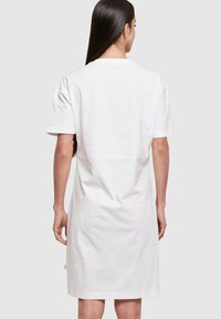Biała bawełniana sukienka t-shirtowa z krótkimi rękawami, luźny krój i prosty design. Posiada widoczną szew z tyłu i małą zieloną metkę.