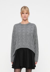 Sivok pulover iz pletenine s kabelskim vzorcem, skrajšane dolžine in širokimi rokavi, v kombinaciji z črno večplastno krilo.