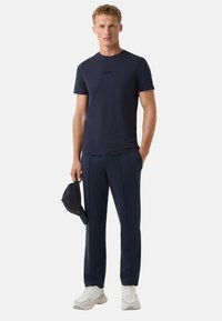 Mørkeblå t-shirt med et diskret logo, parret med matchende mørkeblå bukser og en sort cap i hånden. Sneakers har et robust design med grå detaljer.
