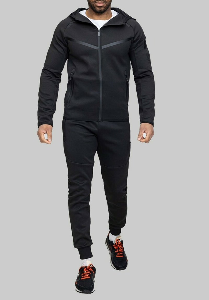 Survêtement noir de sport avec capuche, doté de poches zippées, d'un tissu texturé et de détails contrastés. Associé à des baskets noires avec des lacets orange.