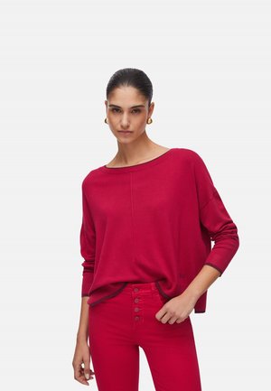 Donna che indossa un maglione rosso ampio con maniche lunghe e pantaloni rossi a vita alta con bottoni, in piedi davanti a uno sfondo bianco.