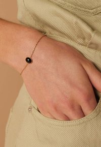 Gold chain bracelet featuring an oval black stone centerpiece, set in a smooth bezel, worn on a hand against a textured fabric backdrop.
