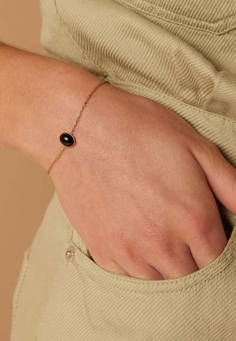 Gold chain bracelet featuring an oval black stone centerpiece, set in a smooth bezel, worn on a hand against a textured fabric backdrop.