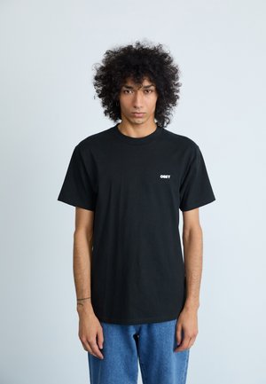 T-shirt in cotone nero con scollo rotondo e maniche corte, con logo bianco "OBEY" sul lato sinistro del petto. Indossato con jeans blu.