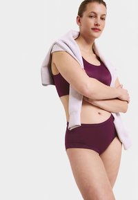 Reggiseno sportivo color bordeaux e slip a vita alta realizzati in tessuto liscio, abbinati a una felpa grigio chiaro drappeggiata sulle spalle.