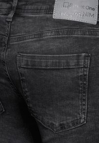 Svarta denimjeans med fem fickor, kontrasterande sömmar och en tygetikett på midjan. Texturen är jämn med en lätt lyster.