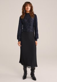 Marinblå långärmad topp med texturerad blommönster, kombinerad med en plisserad svart midikjol och knähöga svarta stövlar.