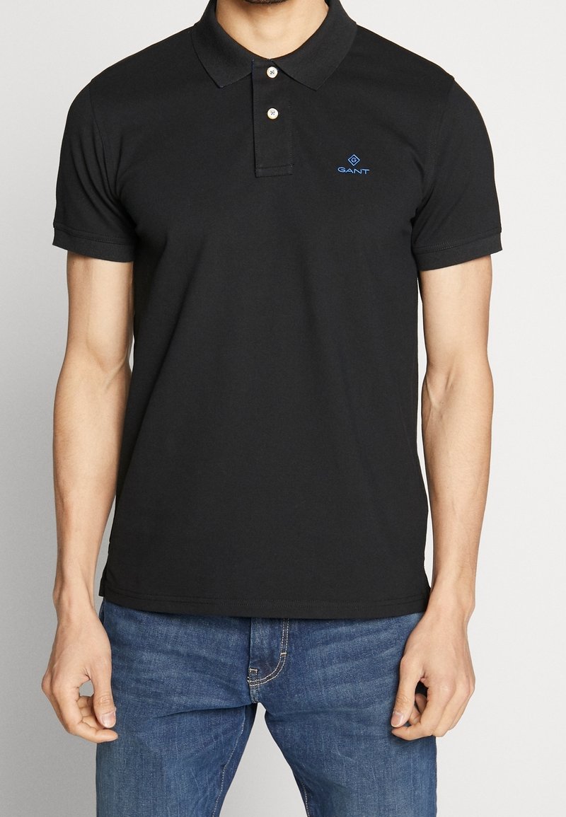 Homme portant un polo noir à manches courtes avec deux boutons et logo bleu GANT, associé à un jean bleu, sur un fond uni.