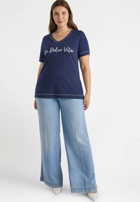 Camiseta de cuello en V azul marino con el texto "La Dolce Vita" en blanco, combinada con unos pantalones vaqueros de pierna ancha en azul claro y zapatos blancos. Textura suave.