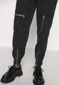 Pantalon noir avec plusieurs fermetures éclair visibles sur les jambes, associé à des chaussures à plateforme épaisses noires à lacets sur un fond clair.