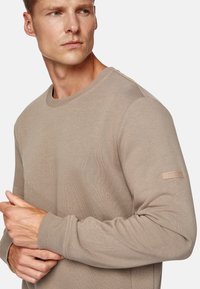 Beige crew neck sweatshirt with ribbed cuffs and hem, featuring a textured finish and a subtle branded patch on the left sleeve.