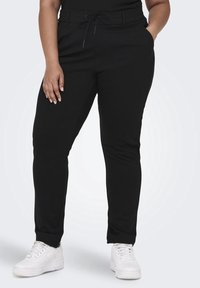 Person som bär svarta smala byxor med dragsko i midjan och vita sneakers, med en hand i fickan, stående mot vit bakgrund.