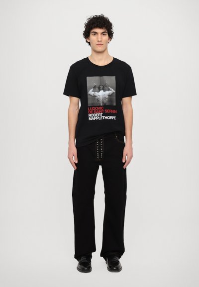 Black t-shirt featuring a graphic print of a muscular figure with text in red and white. Paired with black pants and shiny shoes.