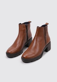 Botas de cuero marrón hasta el tobillo con una textura suave, cremalleras laterales y tachuelas decorativas. Tacón negro grueso y diseño de punta redonda.
