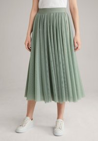 Vaaleanvihreä tylli-hame, jossa on poimutettu malli, joustava vyötärö, jossa on brändin nimi, käytetään valkoisten tennarien kanssa.