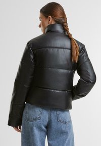 Svart boblejakke laget av glatt syntetisk materiale, kort design med høy krage, med horisontale sømmer og vide ermer.
