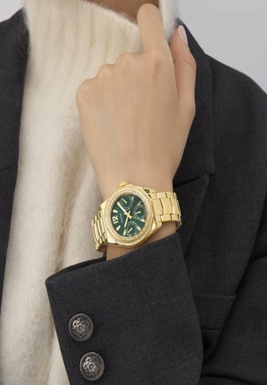 Orologio Guess d'oro con quadrante verde e tre sottocronografi indossato al polso, abbinato a un blazer scuro e un maglione bianco a trama.