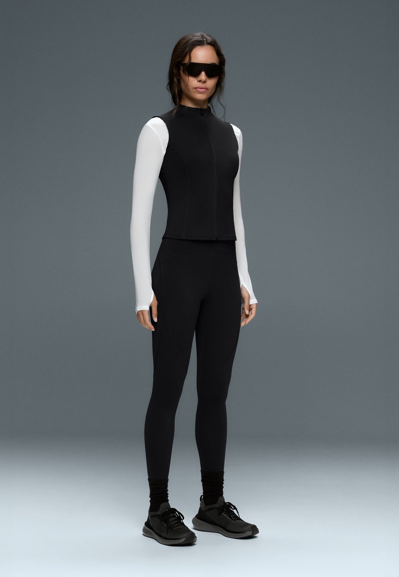 Woman wearing black sleeveless vest over white long-sleeve shirt, black leggings, black sneakers, and dark sunglasses against gray background.