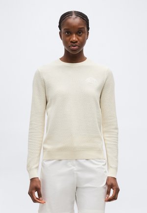 Žena s vrkočmi, oblečená v krémovom pletenom svetri s dlhým rukávom a bielymi nohavicami, stojaca pred jednoduchým svetlým pozadím.