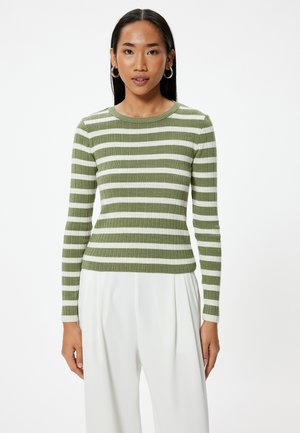 Haut à rayures vertes et blanches, à manches longues et à col rond ; design ajusté ; tissu texturé ; porté avec un pantalon blanc.