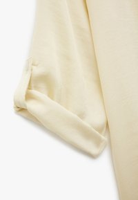 Cremfarbene Bluse mit einer weichen, glatten Textur. Der hochgerollte Ärmel verfügt über einen Knopfverschluss und dezente Stichdetails.
