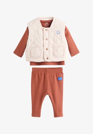 Beige, gesteppte ärmellose Weste mit Knöpfen über rostfarbenem Langarmshirt und passender Hose mit „lächle, kleines“ Aufnäher.