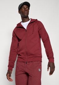 Burgunderfarbener Kapuzenpullover mit Reißverschluss, Zugkordel, Fronttaschen und kontrastierendem weißen Logo auf der linken Brust; kombiniert mit passenden Hosen, beide aus weichem Stoff.