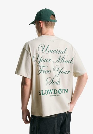 Beige bomulds t-shirt med grøn, kursiv tekst på ryggen. Kappen er grøn med et enkelt design. Modellen har mørke jeans på. Synlig logo: SLOWDØN.