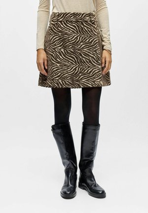Donna con gonna a stampa zebra marrone e beige, collant neri, stivali neri alti fino al ginocchio e top a maniche lunghe beige in piedi contro uno sfondo bianco.