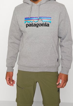 Muž nosí šedou mikinu Patagonia s barevným horským logem a olivově zelené šortky se zipovou kapsou na stehně.