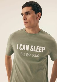 Oliwkowa bawełniana koszulka z okrągłym dekoltem, z wyraźnym białym napisem "MOGĘ SPAĆ CAŁY DZIEŃ". Krótkie rękawy.