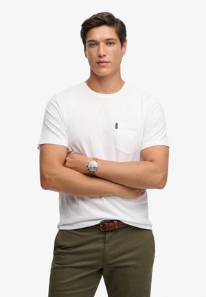 Ung mand med korslagte arme iført hvid t-shirt med lomme, olivengrønne bukser, fletbælte i brun og guldt ur mod hvid baggrund.