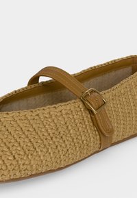 Scarpa piatta intrecciata color cuoio da donna con cinturino in pelle e fibbia dorata sul collo del piede, fodera neutra in tessuto e suola piatta.