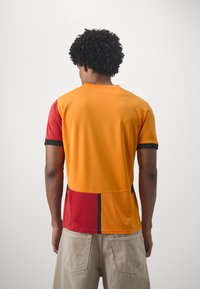 Kortærmet skjorte med orange og røde farveblokke samt sorte detaljer. Stoffet virker let og har en glat tekstur.