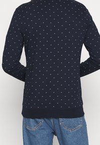 Sudadera azul marino con un pequeño patrón geométrico repetido. Mangas largas, puños y dobladillo acanalados. Llevada sobre unos jeans de denim azul.