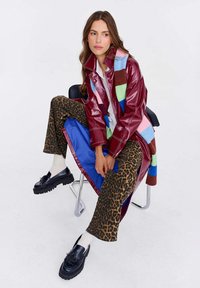 Bordový kabát z faux kože s farebnými vzorovanými akcentmi, v kombinácii s nohavicami v leopardom vzore a čiernymi robustnými mokasínami, sediac na stoličke.